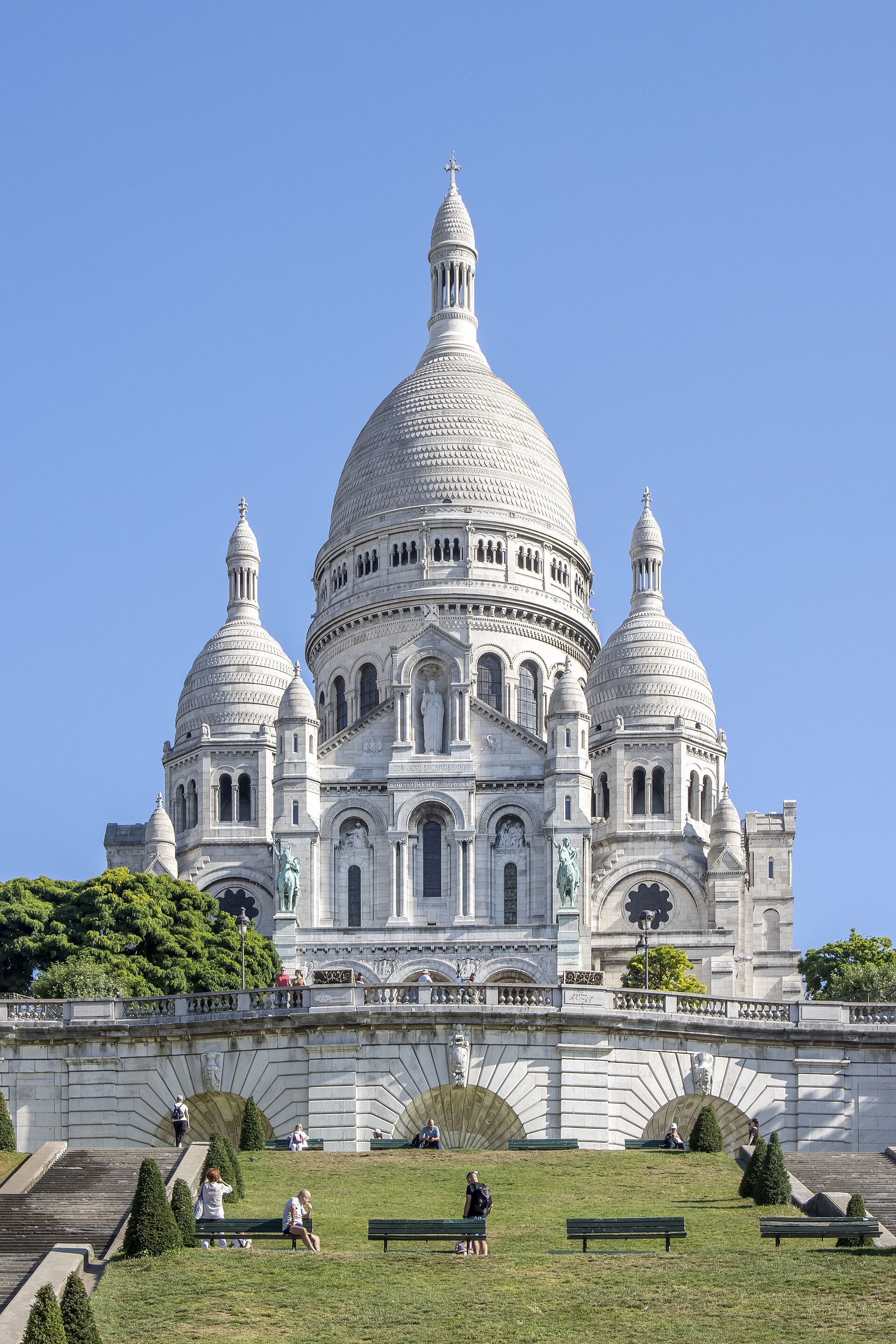 Basilique du Sacré-Cœur à Montmartre, dômes blancs, pelouse, bancs et visiteur au premier plan.