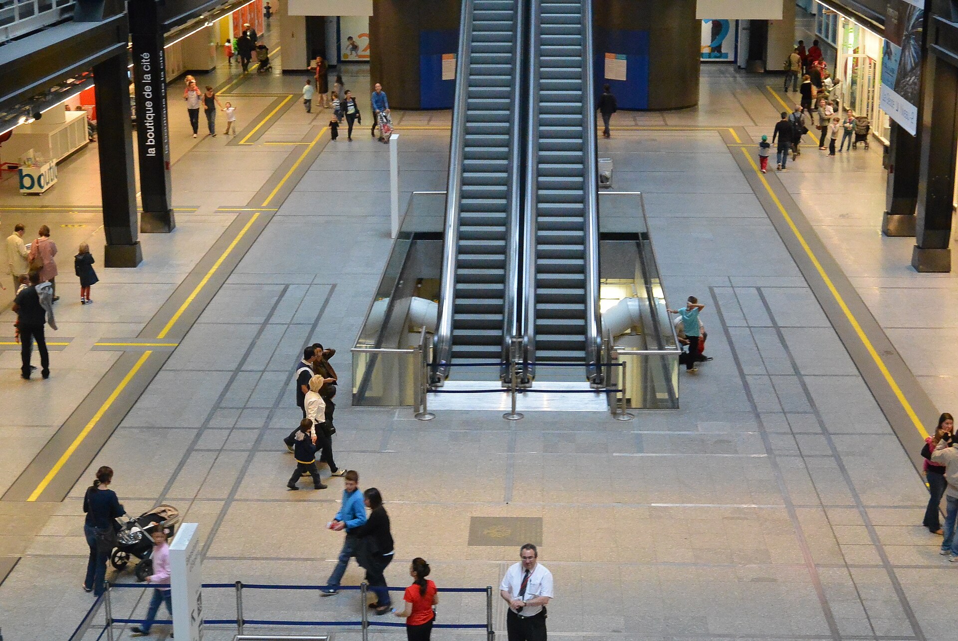 Hall de la Cité des sciences : escalator central, sol marqué de bandes jaunes, public et boutique de la Cité.